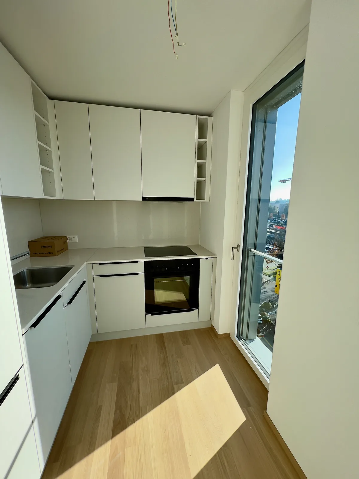 Man sieht eine Kochnische. Links das Spülbecken, Einbauschränke, vorne der Herd und rechts ein bodenlanges Fenster.